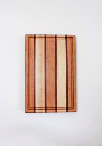 Large cutting board with strips of cherry, purpleheart and maple wood featuring a juice groove.